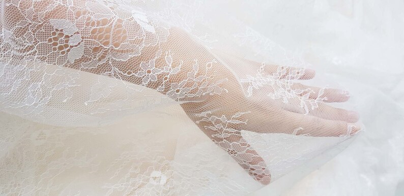May include: White lace fabric with a delicate floral pattern. The fabric is sheer and has a soft, flowing texture.