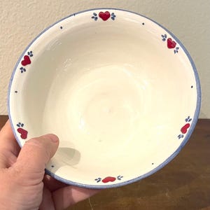 May include: A round ceramic bowl with a cream-colored interior and exterior. The rim is painted with a light blue band, accented by small red heart motifs and tiny blue dots spaced evenly around the circumference of the bowl's edge.