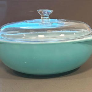 May include: A round, turquoise-colored casserole dish with a smooth, matte finish and integrated side handles. The dish is topped with a clear, circular glass lid that features a small, centered knob handle for lifting and a slightly raised rim.