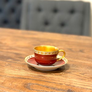 May include: A deep red teacup and matching saucer set featuring a metallic gold interior and a decorative white and gold band around the rim. The cup has a curved handle and sits centered on the circular saucer, showcasing a smooth, polished finish.