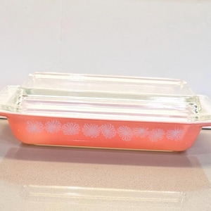 May include: A rectangular pink glass baking dish with a clear glass lid. The exterior of the dish features a repeating white floral pattern along the sides. The dish has integrated handles on both ends and a smooth, glossy finish throughout.