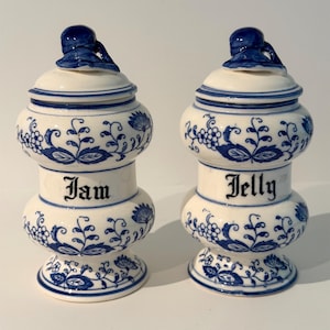 May include: Two white ceramic jars with blue floral patterns and matching lids. One jar is labeled Jam and the other is labeled Jelly in black script. Each jar features a bulbous body, a flared base, and a decorative finial on the lid.