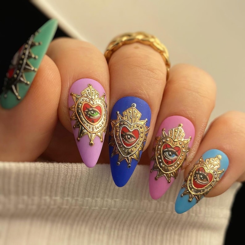 Pode incluir: Cinco unhas compridas em forma de am&ecirc;ndoa s&atilde;o pintadas em cores diferentes e decoradas com pingentes dourados em forma de cora&ccedil;&atilde;o com um design de olho. As unhas s&atilde;o pintadas em tons de rosa, azul e verde.