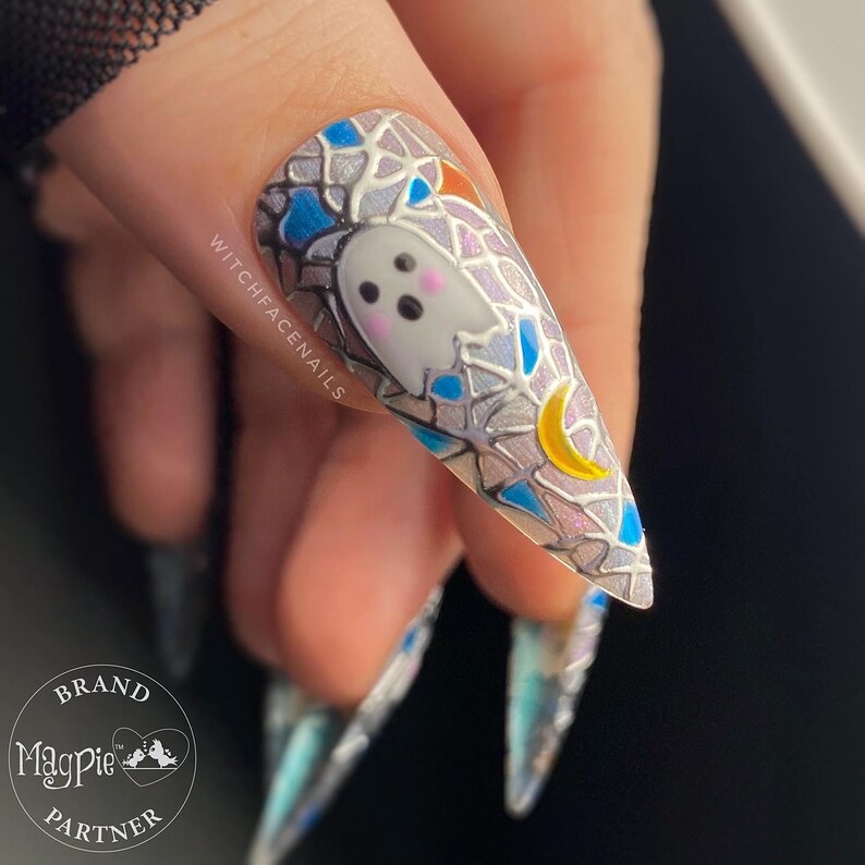 Op de afbeelding: Close-up van een lange, puntige, lichtblauwe en zilveren nagel met een wit spook en een gele halve maan. De nagel is versierd met een glas-in-lood effect. De tekst "WITCHFACENAILS" is zichtbaar op de nagel.