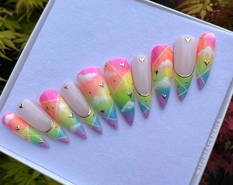 May include: Rainbow ombre press-on nails with white clouds, gold hearts, and a gold accent line. The nails are in a coffin shape and are displayed on a white background. The text "WITCHFACENAILS" is visible in the bottom right corner of the image.