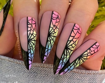 May include: Close-up of long, pointed, nude-colored fingernails with a black spiderweb design and a rainbow ombre effect. The nails are decorated with a black outline and a black spiderweb design.