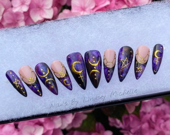 May include: A set of press-on nails with a purple and gold celestial design. The nails feature a mix of solid purple, nude, and patterned designs with moons, stars, and other mystical symbols. The text "Nails By Lindsey Michelle" is visible.