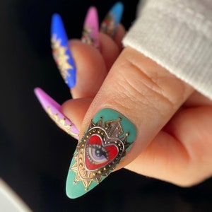 Pode incluir: Close-up de uma unha turquesa com um design em forma de cora&ccedil;&atilde;o dourado que apresenta um cora&ccedil;&atilde;o vermelho com um olho e uma coroa. A arte de unhas &eacute; decorada com glitter dourado.
