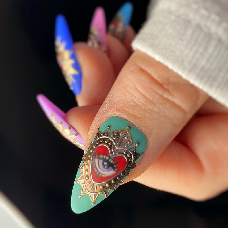 Pode incluir: Close-up de uma unha turquesa com um design em forma de cora&ccedil;&atilde;o dourado que apresenta um cora&ccedil;&atilde;o vermelho com um olho e uma coroa. A arte de unhas &eacute; decorada com glitter dourado.