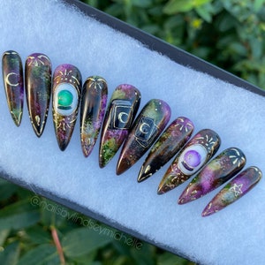 May include: Set of ten long, pointed false nails with a black base and a galaxy-like design. The nails are decorated with gold accents, green and purple gemstones, and various symbols, including a crescent moon, a pentagram, and a tarot card.