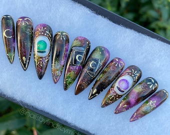 May include: Set of ten long, pointed false nails with a black base and a galaxy-like design. The nails are decorated with gold accents, green and purple gemstones, and various symbols, including a crescent moon, a pentagram, and a tarot card.