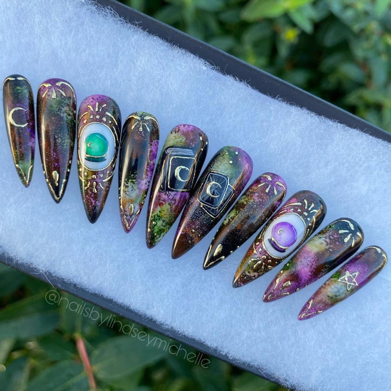 May include: Set of ten long, pointed false nails with a black base and a galaxy-like design. The nails are decorated with gold accents, green and purple gemstones, and various symbols, including a crescent moon, a pentagram, and a tarot card.