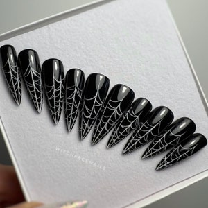 May include: Black, glossy, coffin-shaped press-on nails with a white spiderweb design. The nails are arranged in a white box. The text "WITCHFACENAILS" is printed on the box.