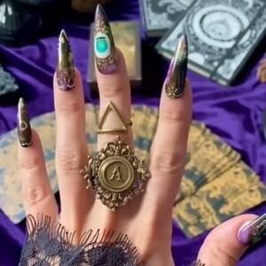 May include: A hand with long, pointed, black and gold painted nails, wearing a gold ring with an ornate design and a letter 'A' in the center. The nails are decorated with various designs, including an eye with a green gemstone.
