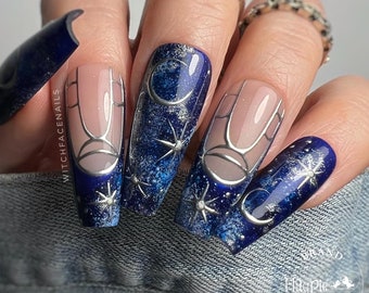 May include: Close-up of five fingernails with a dark blue and silver galaxy design. The nails are long and almond shaped. The design features silver crescent moons, stars, and swirls. The text "WitchfaceNails" is visible on one of the nails.