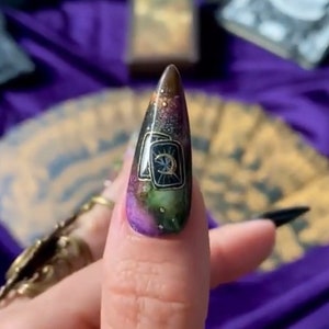 May include: A long, pointed, black nail with a purple, green, and gold galaxy design. The design features a tarot card with a crescent moon and stars.