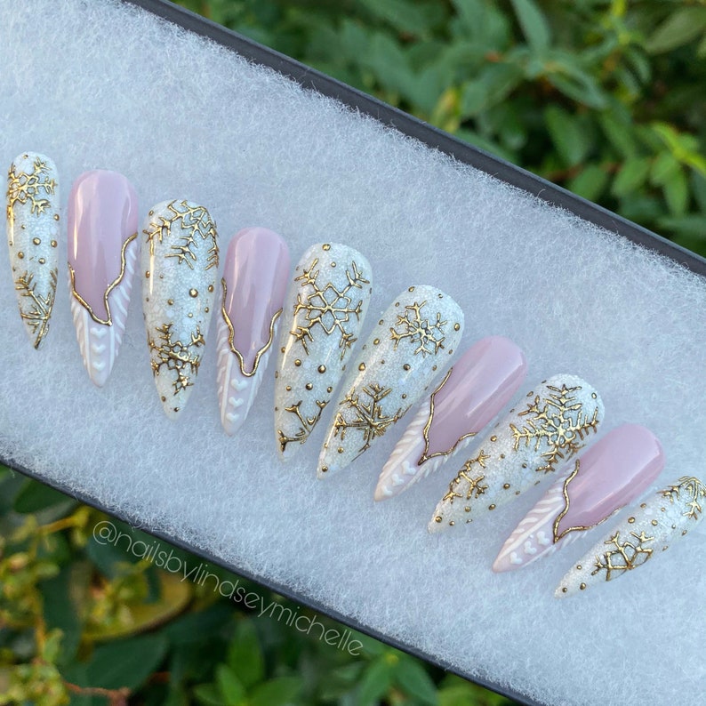 May include: Set of ten white and pink stiletto false nails with gold snowflake designs and white accents. The nails are arranged in a row on a white surface. @nailsbylindseymichelle