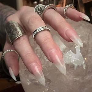 May include: A hand with long, pointed, ombre-style fingernails, transitioning from a pink base to a white tip. Several silver rings adorn the fingers, including a band with clear stones. The text "WITCHFACENAILS" is visible.