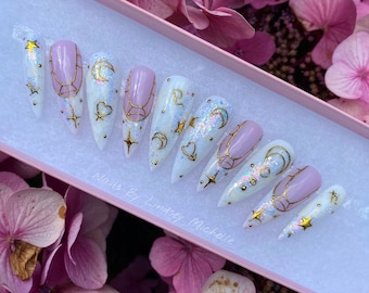 May include: A set of long, almond-shaped press-on nails. The nails feature a white base with gold celestial designs, including stars, moons, and hearts. Some nails have a light pink polish. The box is light pink, and the text "Nails By Lindsey Michelle" is visible.
