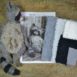 Può includere: Un peluche di procione fatto a mano con una coda a strisce nere e grigie e una maschera nera intorno agli occhi. Il peluche è accanto a una foto in bianco e nero di un procione e campioni di tessuto. Il testo "Geralt 23 cm / 9 in" è visibile.