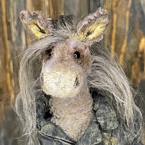 May include: A handmade plush donkey toy with long gray hair, wearing a gray denim jacket with silver buttons. The donkey has a brown face with a white nose and black eyes.