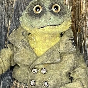 Puede incluir: Una rana de peluche que lleva una chaqueta verde con botones y pantalones a cuadros. La rana tiene ojos grandes y redondos y una expresión amigable.