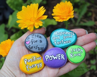 May include: Five painted rocks with inspirational messages. The rocks are painted in various colors, including gray, blue, yellow, purple, and green. The messages on the rocks include "a little for a Grey Day", "Happiness and good fortune", "Positive Energy", "POWER", and "Lucky Rock".