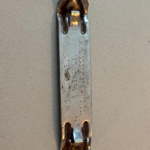 May include: A vintage metal can opener with a pointed end and a rectangular handle. The handle has the text "M.K. Goetz Brewing Co. St. Joseph & Kansas City, MO." The can opener is silver with some areas of rust.