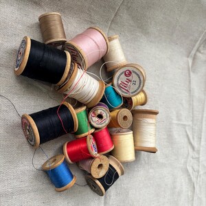 May include: A collection of assorted sewing thread spools in various colors including black, white, red, blue, pink, green, and yellow. The spools are made of wood or plastic and feature labels with text such as Lily and Coats, with loose thread ends.