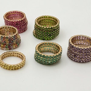 Könnte beinhalten: Eine Sammlung goldfarbener Ringe, die jeweils mit farbigen Edelsteinen besetzt sind. Die Ringe weisen eine Vielzahl von Farben auf, darunter Rosa, Grün, Lila, Gelb und eine Mischung aus anderen. Die Edelsteine sind eng aneinander gesetzt und bilden ein durchgehendes Farbband.