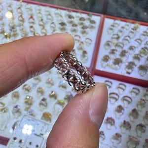May include: A delicate, open-work ring with a repeating geometric pattern. The ring is held between fingers, showcasing its intricate design. The ring appears to be made of a light-colored metal, possibly gold or silver, and is displayed in a jewelry setting.