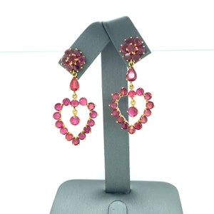 Op de afbeelding: Een paar gouden oorbellen met roze robijnen. De oorbellen zijn hartvormig met een kleiner hart in het midden. De oorbellen hangen aan een klein, bloemvormig cluster van robijnen.