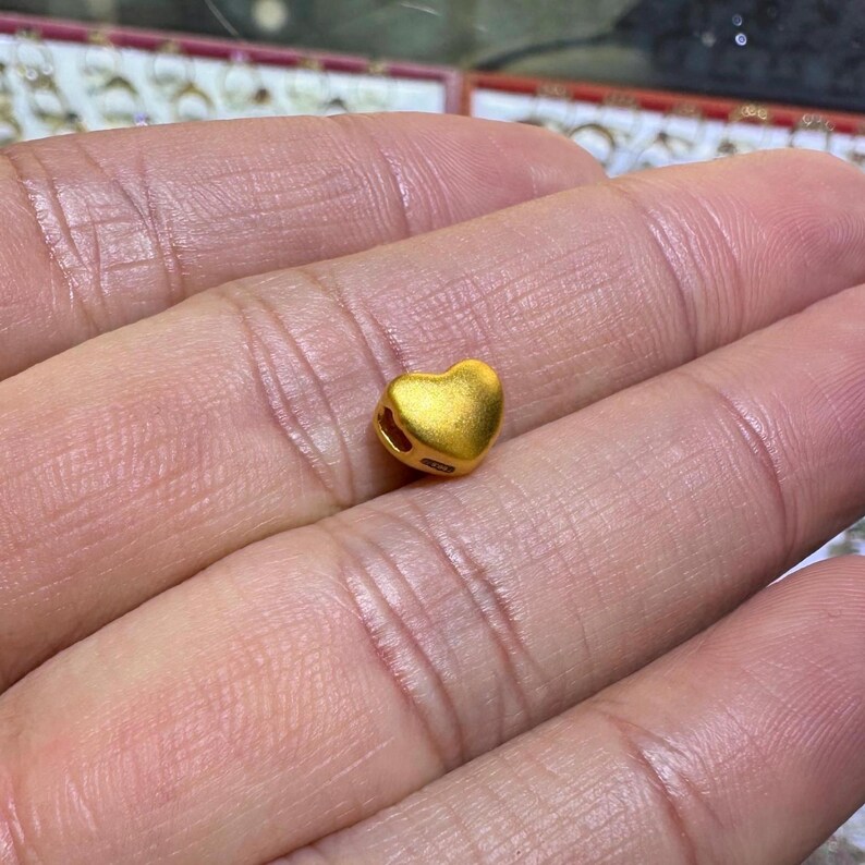 Pode incluir: Uma pequena conta dourada em forma de cora&ccedil;&atilde;o. A conta tem uma superf&iacute;cie lisa e parece ser feita de metal. O cora&ccedil;&atilde;o tem um buraco em cada lado, provavelmente para enfiar. A conta &eacute; segurada na m&atilde;o de uma pessoa.