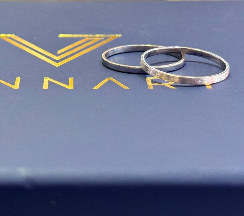 May include: Two silver wedding bands, one slightly larger than the other, are displayed on a blue box with the word "ANNARI" in gold lettering.