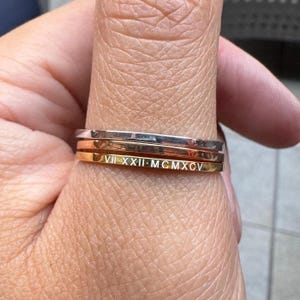 May include: Three stackable rings in silver, rose gold, and gold. The gold ring has Roman numerals etched into it. The rings are simple bands, perfect for everyday wear. The rings are shown on a finger.