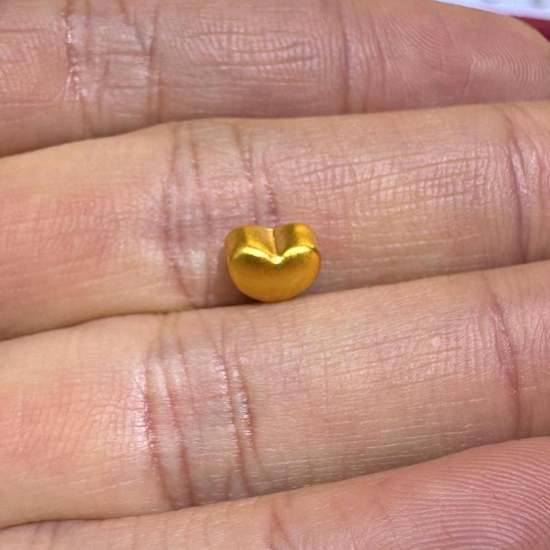 Pode incluir: Um pequeno pingente de ouro em forma de cora&ccedil;&atilde;o. O pingente &eacute; de cor dourada s&oacute;lida e brilhante e tem uma superf&iacute;cie lisa. A forma do cora&ccedil;&atilde;o &eacute; claramente definida, com uma base arredondada e uma ligeira reentr&acirc;ncia no topo. O pingente &eacute; segurado em uma m&atilde;o.