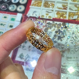 Può includere: Un anello color oro con un motivo traforato ripetuto di cerchi e quadrati ad incastro. L'anello è tenuto contro uno sfondo di altri gioielli.