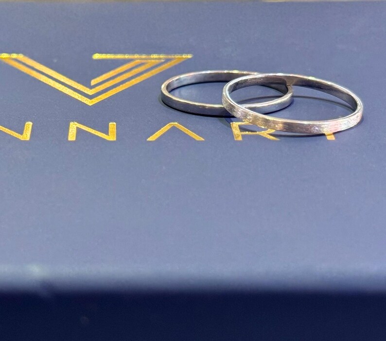 May include: Two silver wedding bands, one slightly larger than the other, are displayed on a blue box with the word "ANNARI" in gold lettering.