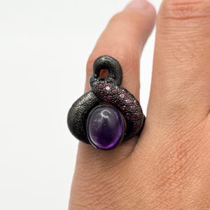 Könnte beinhalten: Ein schwarzer Ring mit einem großen violetten Amethyst-Edelstein und kleineren rosafarbenen Edelsteinen. Der Ring ist in einer einzigartigen, abstrakten Form gestaltet.