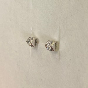 May include: A pair of small, silver-toned stud earrings. Each earring features a geometric design with a clear, sparkling gemstone set in the center. The earrings are displayed against a light background.