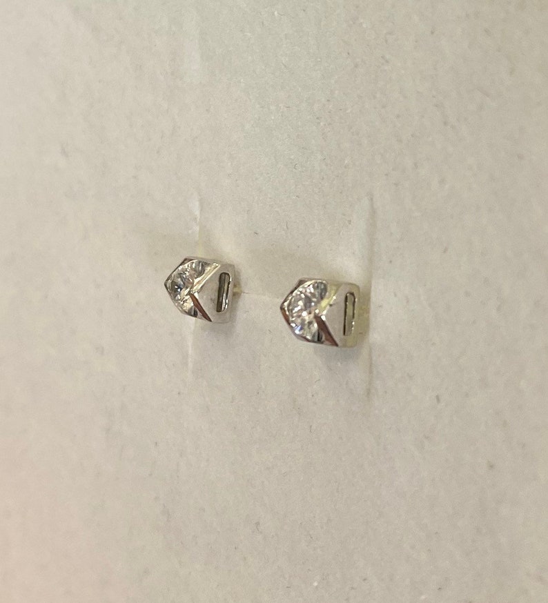 May include: A pair of small, silver-toned stud earrings. Each earring features a geometric design with a clear, sparkling gemstone set in the center. The earrings are displayed against a light background.