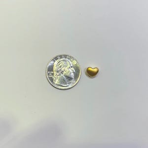 Pode incluir: Um pequeno pingente dourado em forma de cora&ccedil;&atilde;o ao lado de uma moeda de vinte e cinco centavos americana. O pingente &eacute; pequeno, com uma forma lisa e arredondada. A moeda mostra o perfil de George Washington e as palavras "Liberty" e "In God We Trust".