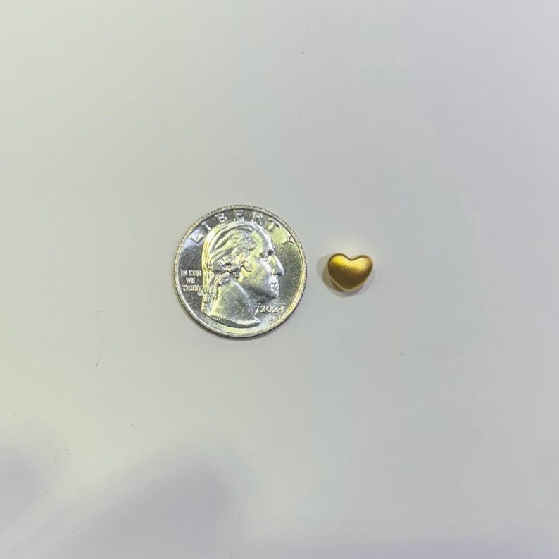Pode incluir: Um pequeno pingente dourado em forma de cora&ccedil;&atilde;o ao lado de uma moeda de vinte e cinco centavos americana. O pingente &eacute; pequeno, com uma forma lisa e arredondada. A moeda mostra o perfil de George Washington e as palavras "Liberty" e "In God We Trust".