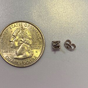 May include: A silver-colored earring stud with a square design and a matching earring back, next to a U.S. quarter coin. The earring stud is small, likely for pierced ears.