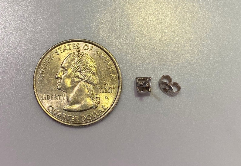 May include: A silver-colored earring stud with a square design and a matching earring back, next to a U.S. quarter coin. The earring stud is small, likely for pierced ears.