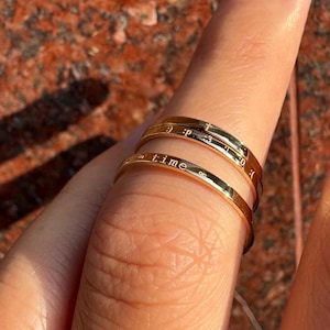 May include: Two gold rings worn on a finger. The top ring has the numbers 1 to 12 engraved on it, and the bottom ring has the word "time" engraved on it.