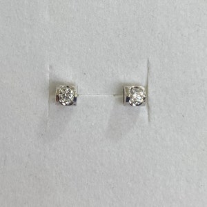 May include: A pair of small, square-shaped stud earrings. Each earring features a single, round-cut diamond set in a silver or white gold bezel. The earrings are displayed on a white surface.