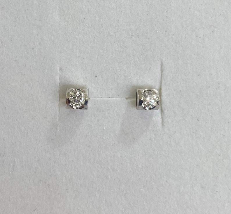May include: A pair of small, square-shaped stud earrings. Each earring features a single, round-cut diamond set in a silver or white gold bezel. The earrings are displayed on a white surface.