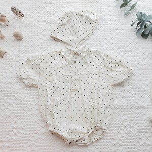 May include: A white baby onesie with black polka dots and a matching white hat with black polka dots. The onesie has a ruffled neckline and short sleeves.