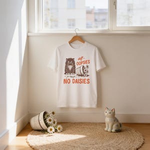 a white t-shirt hanging on a wooden hanger, with a cat figurine and a basket of flowers placed on a rug in front of it.